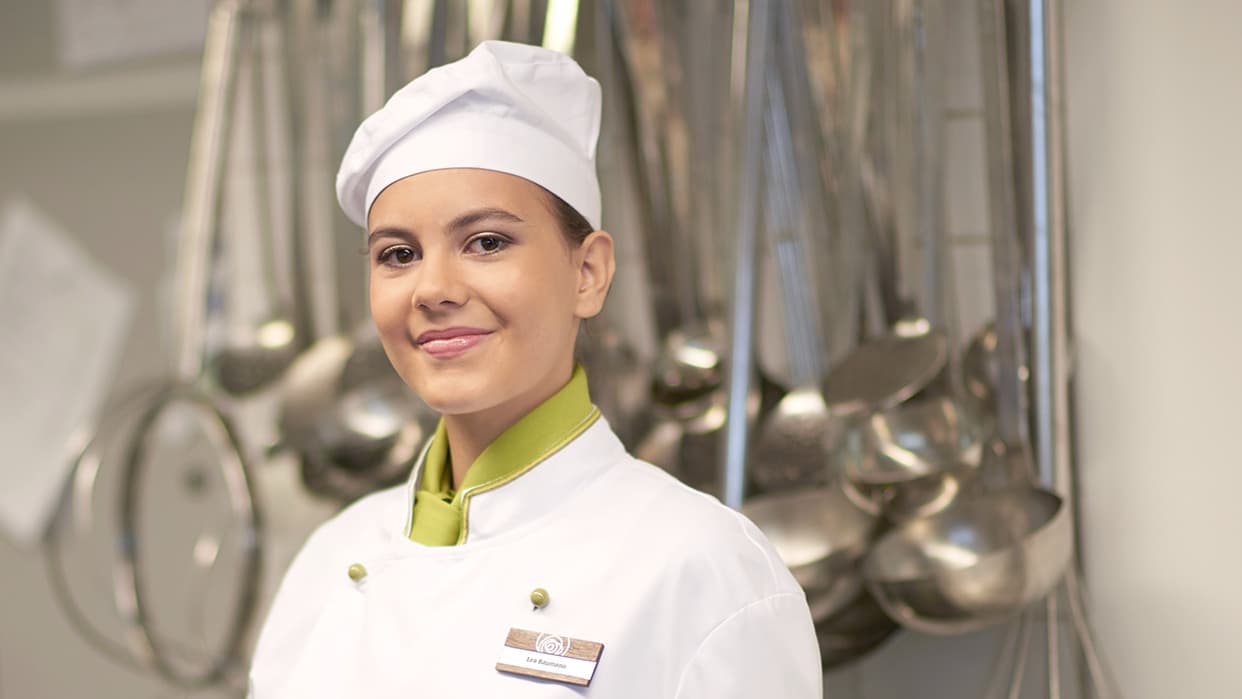Köchin in weisser Uniform in einer Küche mit Kochutensilien im Hintergrund.
