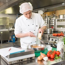 Koch reibt Käse in industrieller Küche mit Zutaten auf dem Tisch.