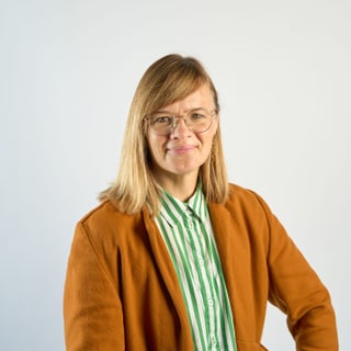 Frau mit Brille und gestreiftem Hemd, trägt einen braunen Mantel, neutraler Hintergrund.