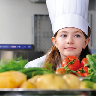 Kind mit Kochmütze in der Küche neben Gemüse und Kartoffeln.