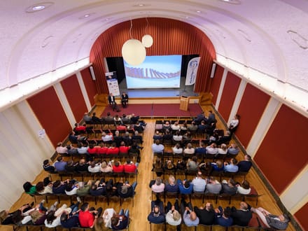 Luftaufnahme eines gefüllten Konferenzraums mit Präsentation auf grosser Leinwand.