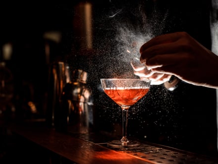 Nahaufnahme eines Cocktails auf einer Theke mit Händen, die Zitruszesten reiben.