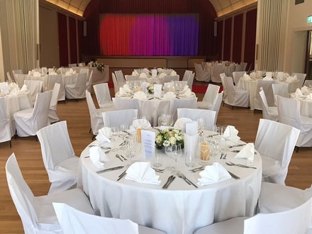 Festlich gedeckte Tische in einem Saal mit Bühne.
