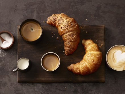 Zwei Croissants und drei Tassen Kaffee auf einem Holzbrett.