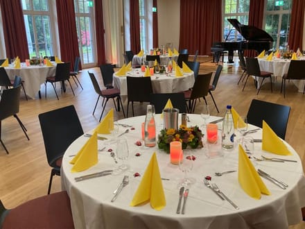 Festlich gedeckter Saal mit runden Tischen und gelben Servietten, Klavier im Hintergrund.
