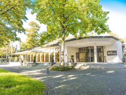 Gebäude mit Bäumen und gepflastertem Vorplatz bei Sonnenschein.