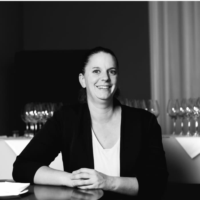 Person in einem Anzug sitzt an einem Tisch mit Weingläsern im Hintergrund.