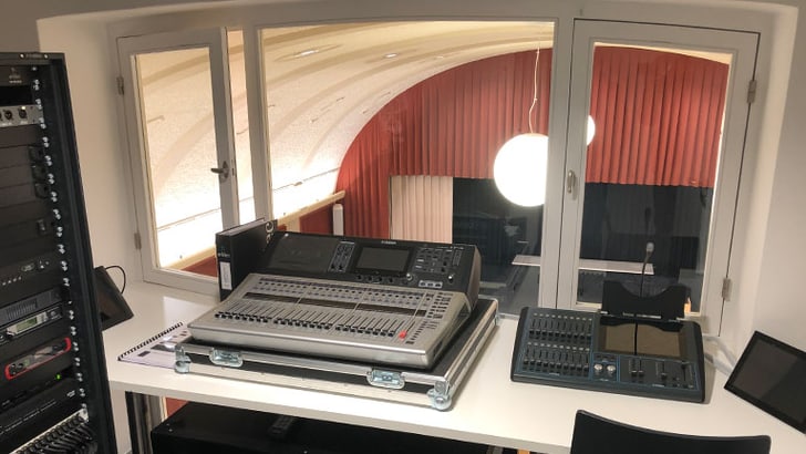 Mischpult vor Fenster, Ausblick auf Bühne mit roten Vorhängen.