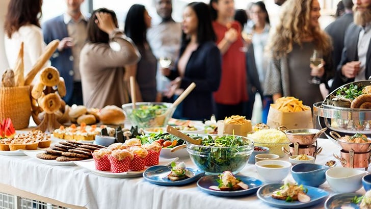 Buffettisch mit verschiedenen Speisen, Menschen im Hintergrund stehend.