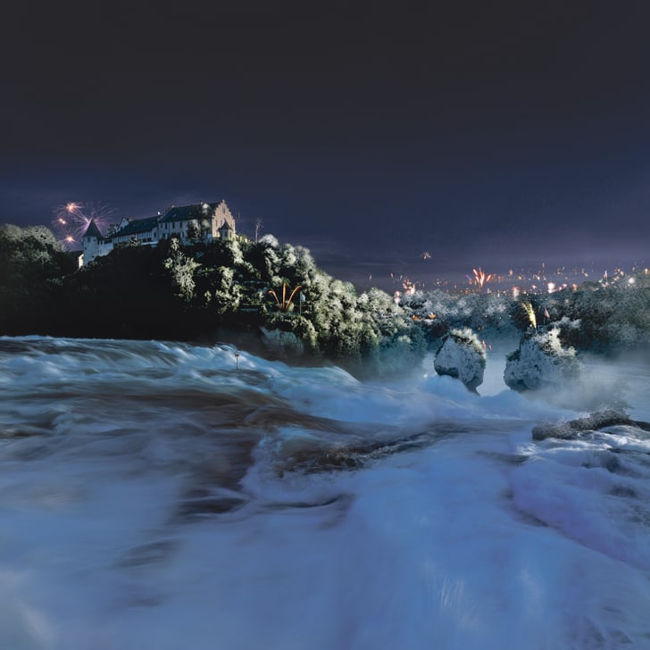 Wasserfall bei Nacht mit Schloss und Feuerwerk im Hintergrund.