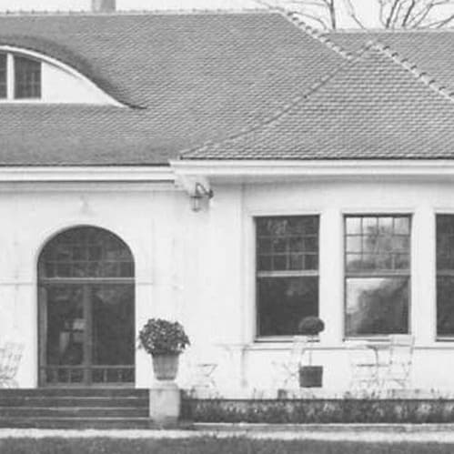 Schwarzweissfoto eines grossen Hauses mit Rundbogenfenstern und Terrassenmöbeln.