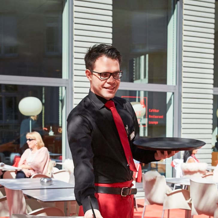 Kellner mit Tablett bedient Gast im Aussenbereich eines Cafés.