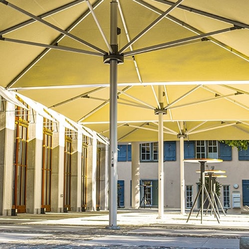 Terrasse mit grossen Sonnenschirmen und Pflanzenkübeln vor einem Gebäude.