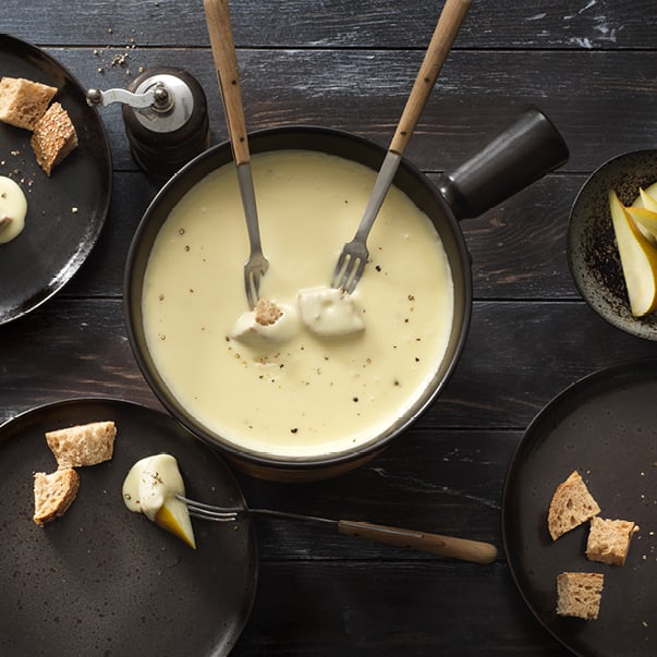Käsefondue mit Brot und Birnen auf dunklem Tisch.