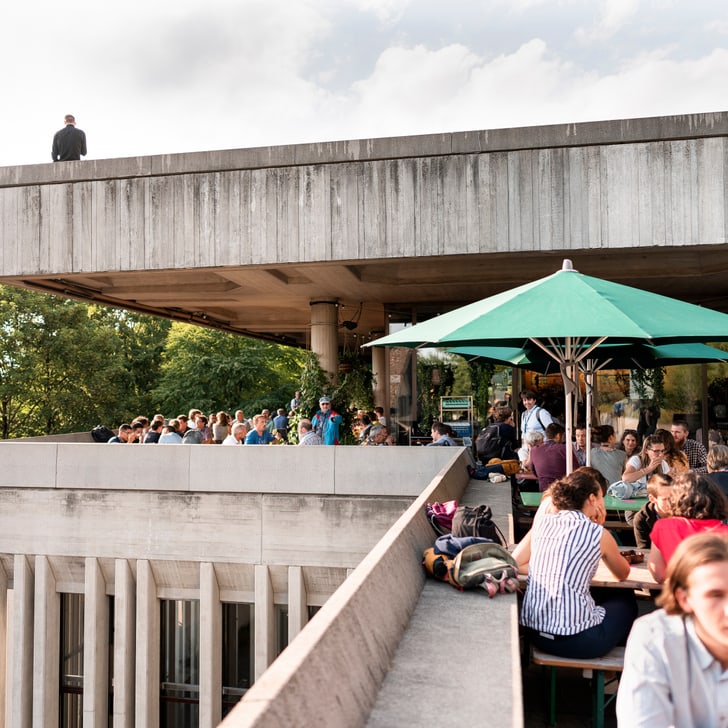 Menschen in einem Café auf einer Dachterrasse bei Sonnenschirmen.",