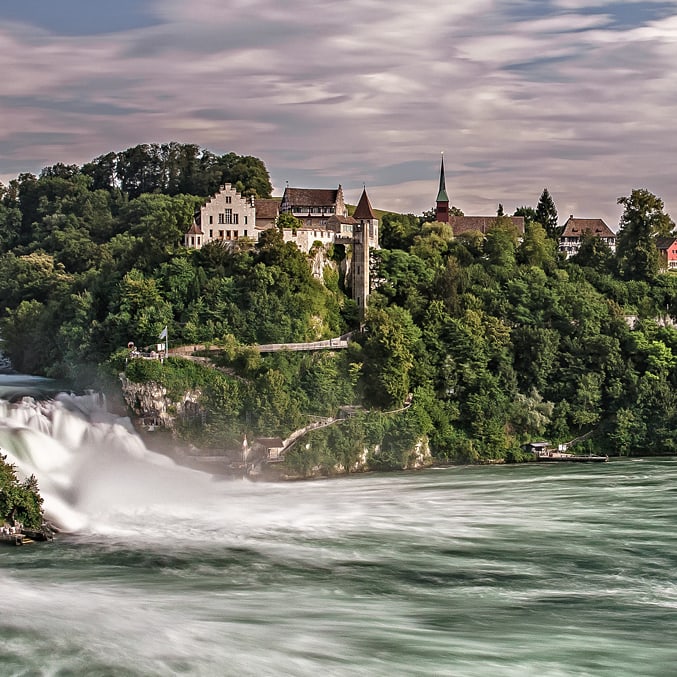 Rheinfall mit Schloss und bewaldetem Ufer.",