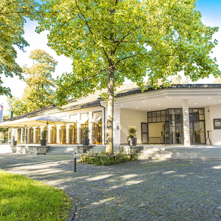 Gebäude mit Bäumen und gepflastertem Vorplatz bei Sonnenschein.