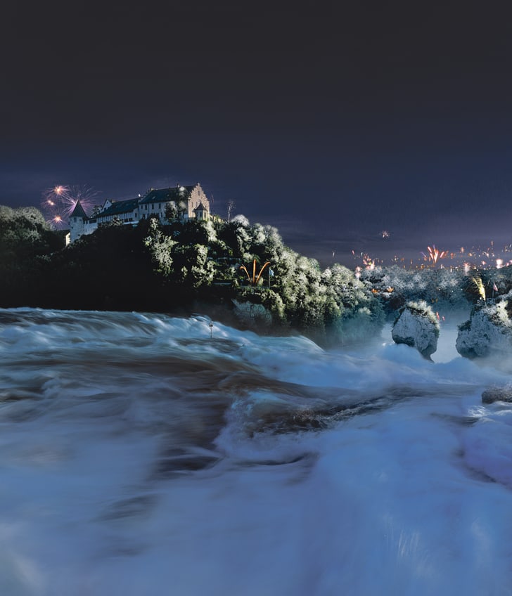 Wasserfall bei Nacht mit Schloss und Feuerwerk im Hintergrund.