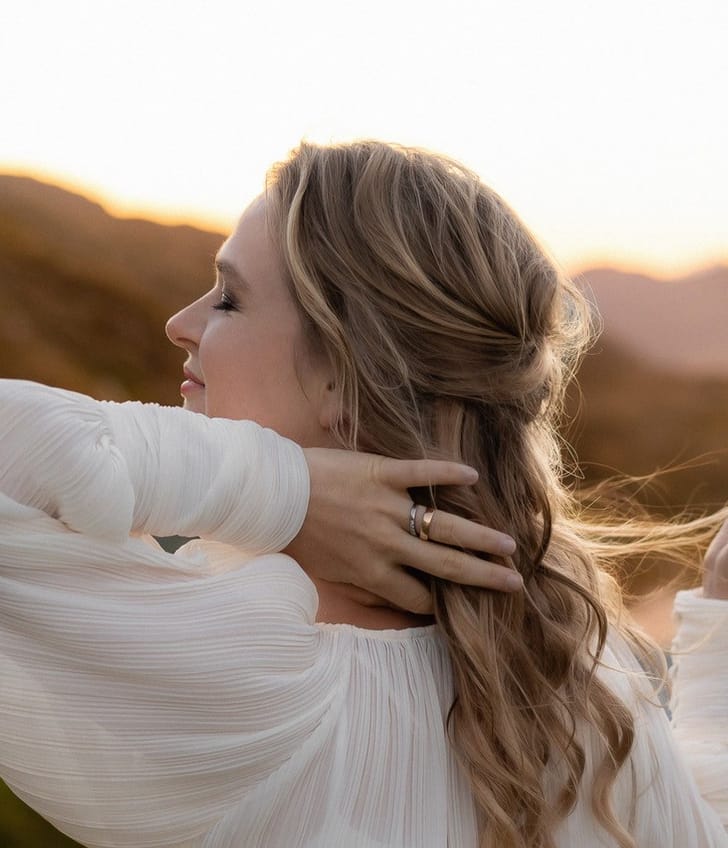 Frau in weissem Kleid vor hügeliger Landschaft.