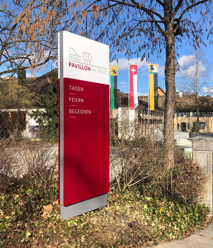 Schild des Pavillons im Park mit Flaggen im Hintergrund.