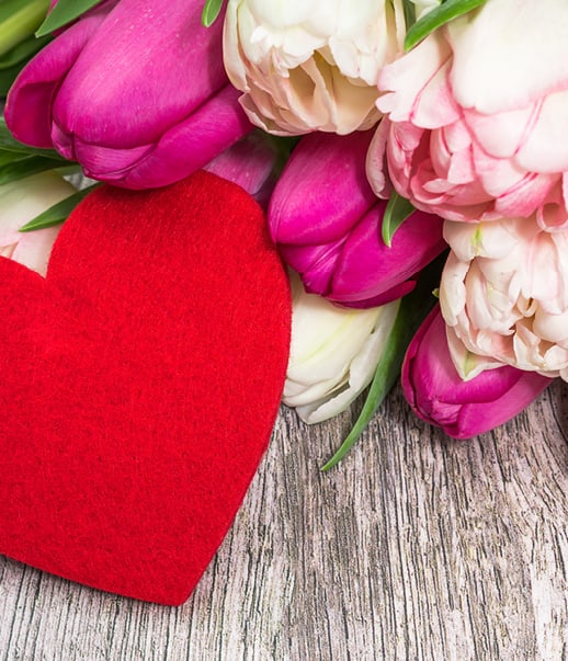 Rotes Herz neben rosa und weissen Tulpen auf Holzoberfläche.