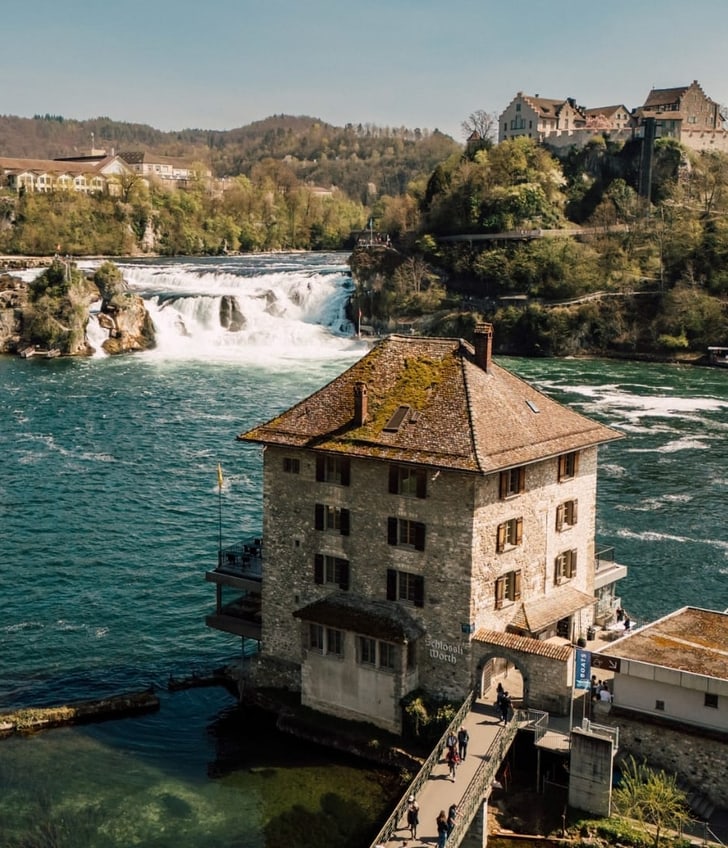 Landschaft mit Fluss, Wasserfall und historischen Gebäuden.