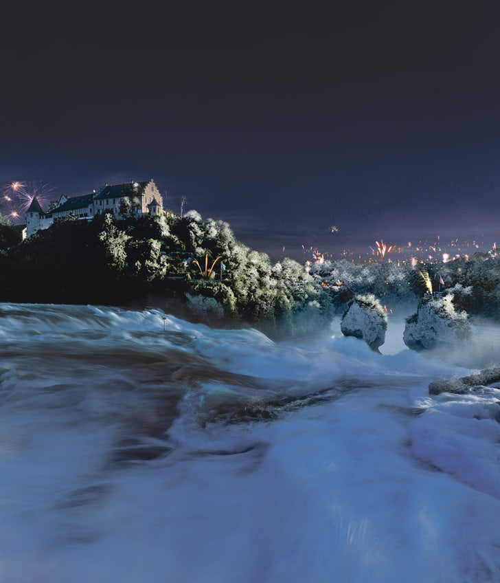 Nachts beleuchteter Wasserfall mit Feuerwerk im Hintergrund und Gebäude auf Hügel.