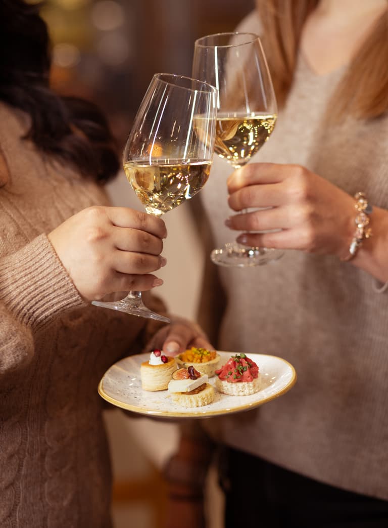 Zwei Personen stossen mit Weingläsern an, halten Teller mit Häppchen.