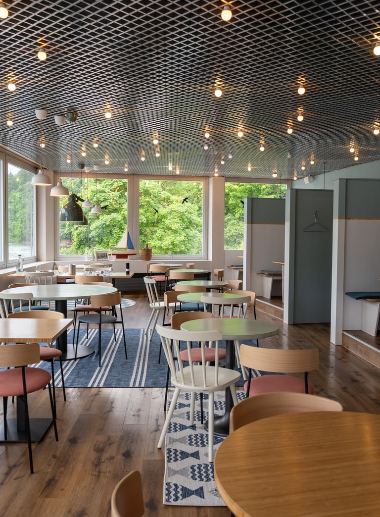 Helles Café mit Holzstühlen und Tischen, grossen Fenstern und Lichterkette an der Decke.