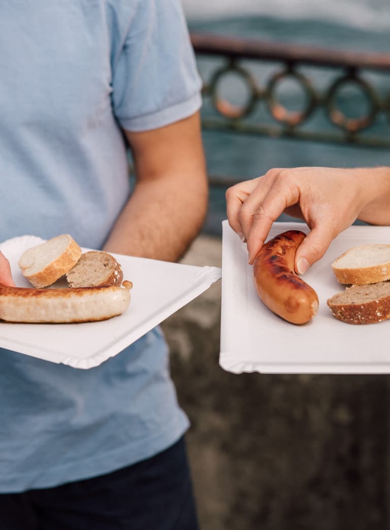 Zwei Personen mit Papptellern und Würstchen im Freien.