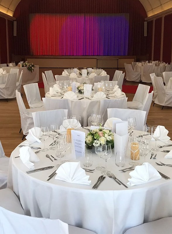 Festlich gedeckte Tische in einem Saal mit Bühne.