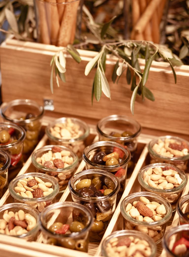 Verschiedene Gläser mit Oliven und Nüssen auf Holztablett.
