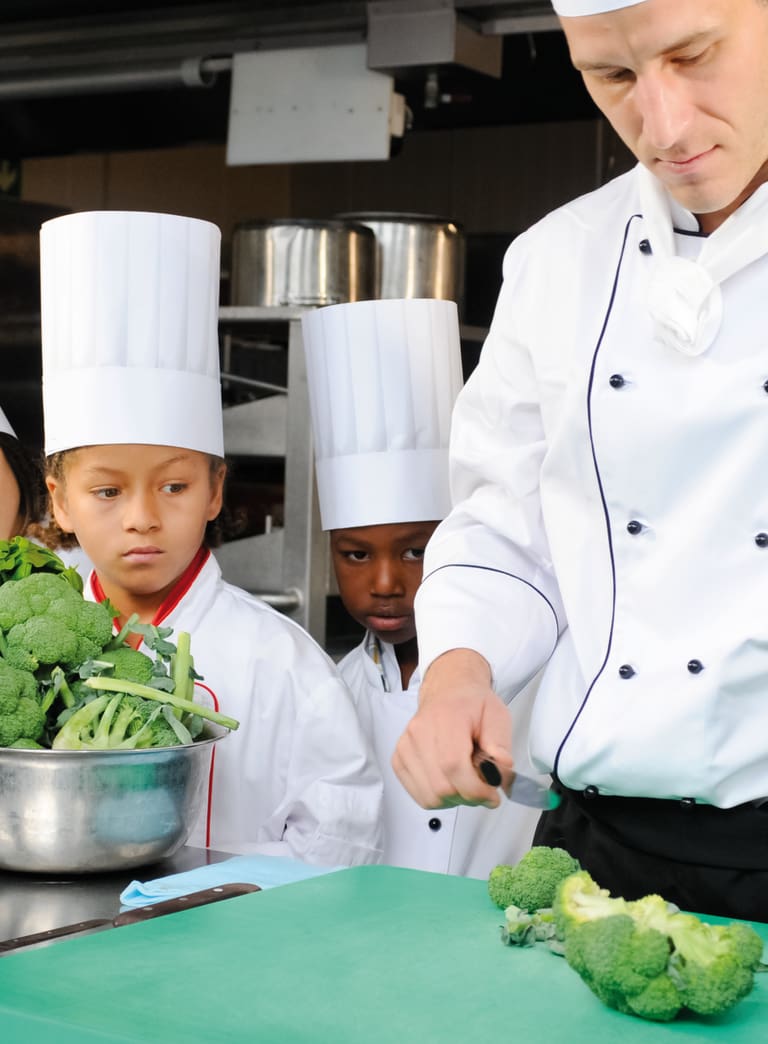 Koch unterrichtet Kinder in einer Küche mit Gemüse auf dem Tisch.",