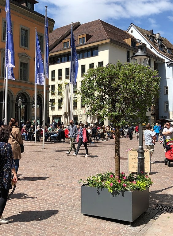 Menschen auf einem belebten Platz mit alten Gebäuden und Fahnen.
