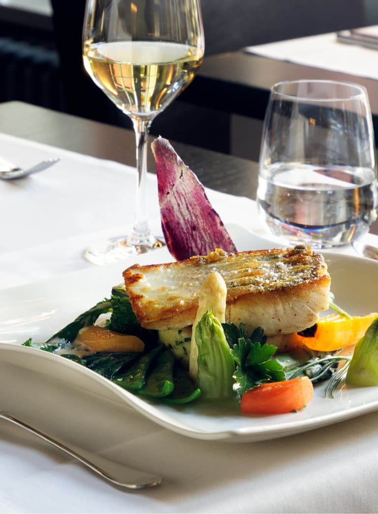 Gourmetgericht mit Fischfilet, Gemüse, Weingläsern auf Tisch eingestellt.