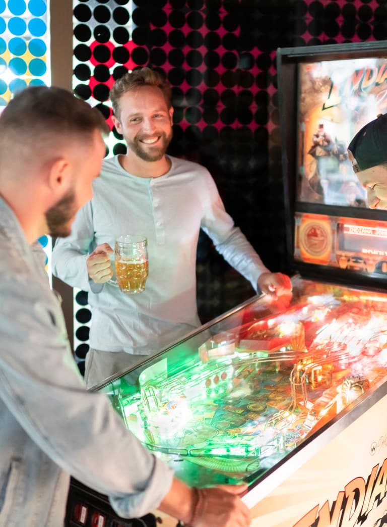 Drei Männer spielen Flipper und halten Bierkrüge.