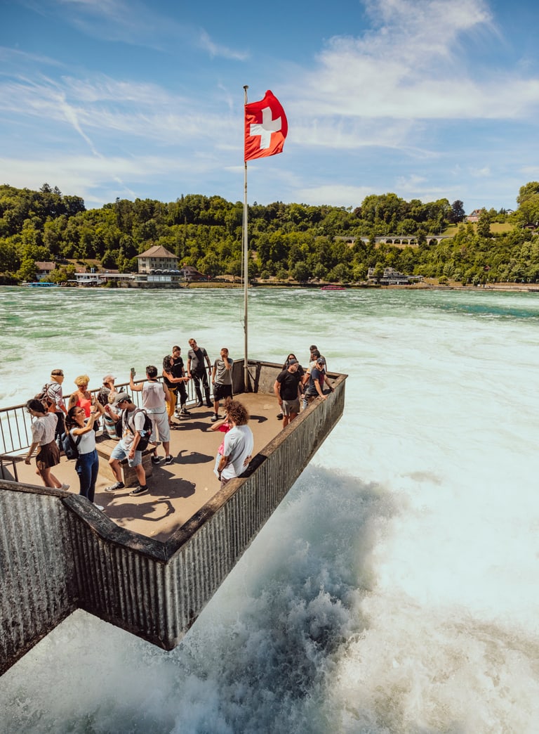 Touristen auf Plattform über Rheinfall mit Schweizer Flagge."