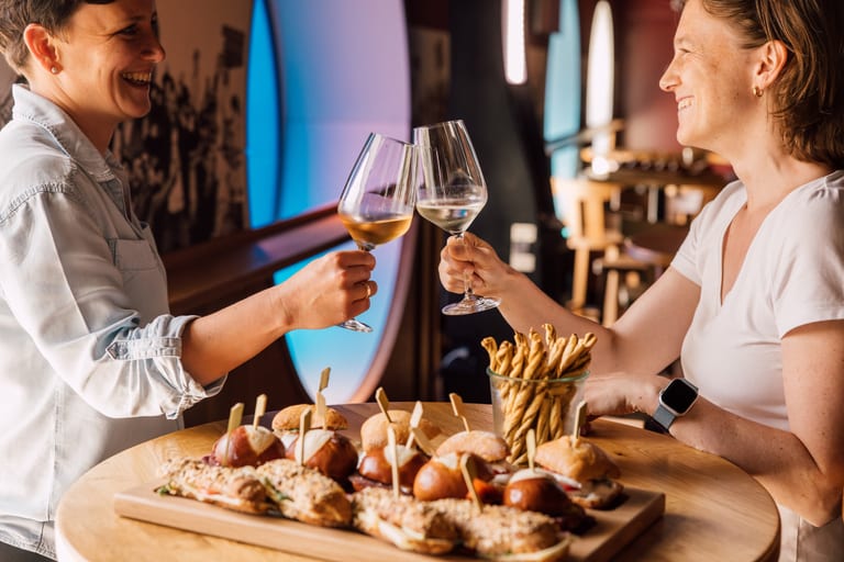 Zwei Personen stossen im Restaurant mit Weingläsern an, Snacks auf dem Tisch.