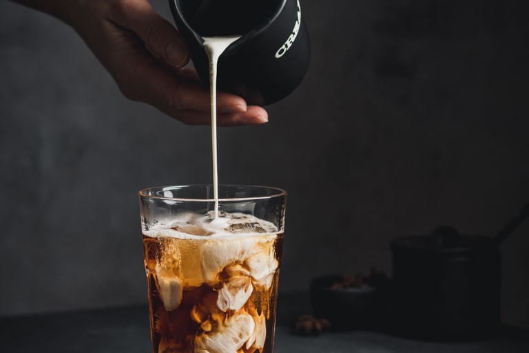 Milch wird in ein Glas Eiskaffee gegossen, auf einem Tisch mit Kaffeebohnen.