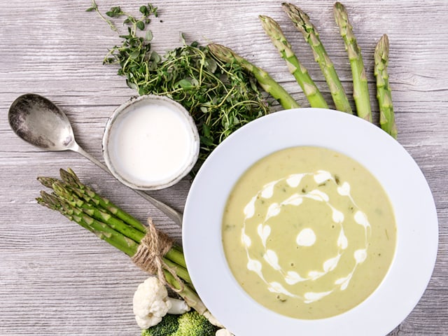 Teller mit grüner Cremesuppe, umgeben von Spargel und Gemüse auf Holzoberfläche.
