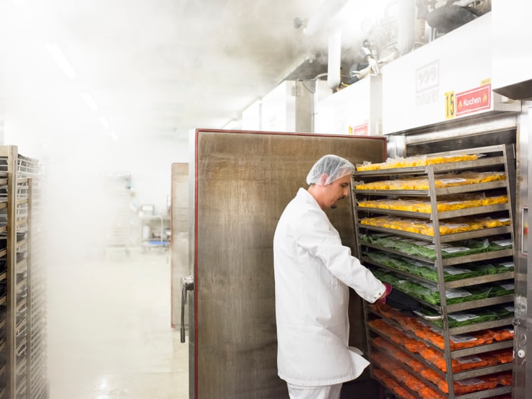 Arbeiter in Lebensmittelverarbeitungsbetrieb mit Dampfschwaden und Tabletts mit Gemüse.
