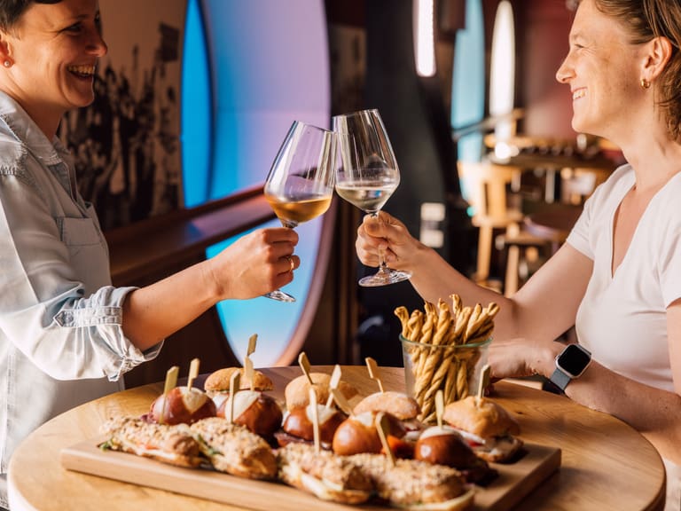 Zwei Personen stossen im Restaurant mit Weingläsern an, Snacks auf dem Tisch.