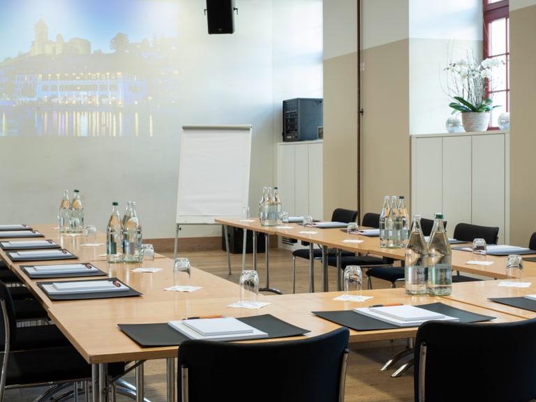 Konferenzraum mit U-förmigen Tischen und leeren Stühlen.