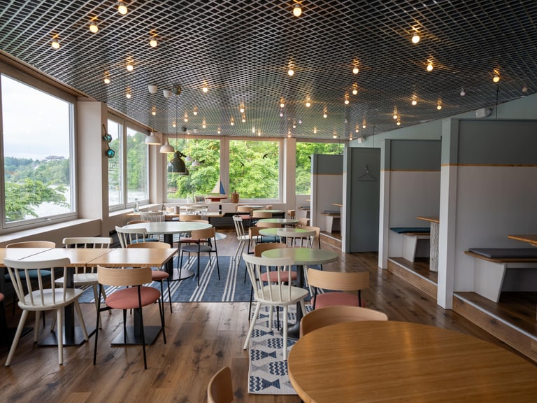 Helles Café mit Holzstühlen und Tischen, grossen Fenstern und Lichterkette an der Decke.