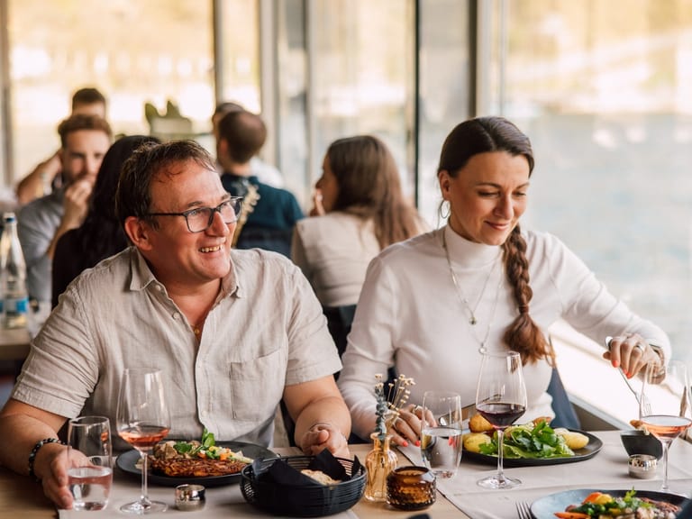 Personen essen lächelnd in einem Restaurant am Wasser.