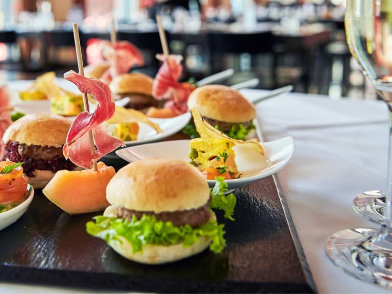 Vorspeisenplatte mit Mini-Burgern und Champagnergläsern auf Tisch.