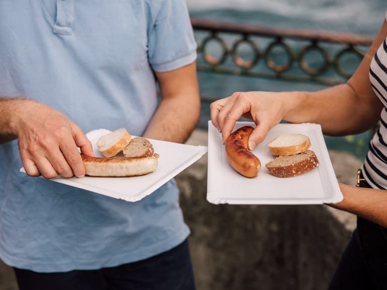 Zwei Personen mit Papptellern und Würstchen im Freien.