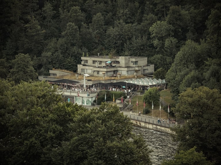 Gebäude von Bäumen umgeben nahe einem Flussufer, mit vielen Menschen davor.