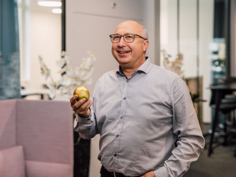Mann mit Brille hält Apfel in der Hand, lächelnd im Büro.