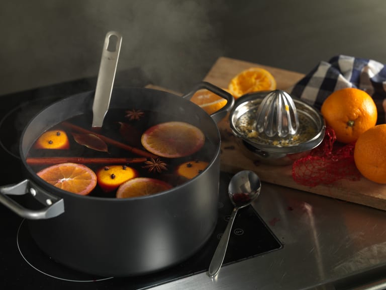 Topf mit Glühwein auf Herd, frische Orangen daneben.
